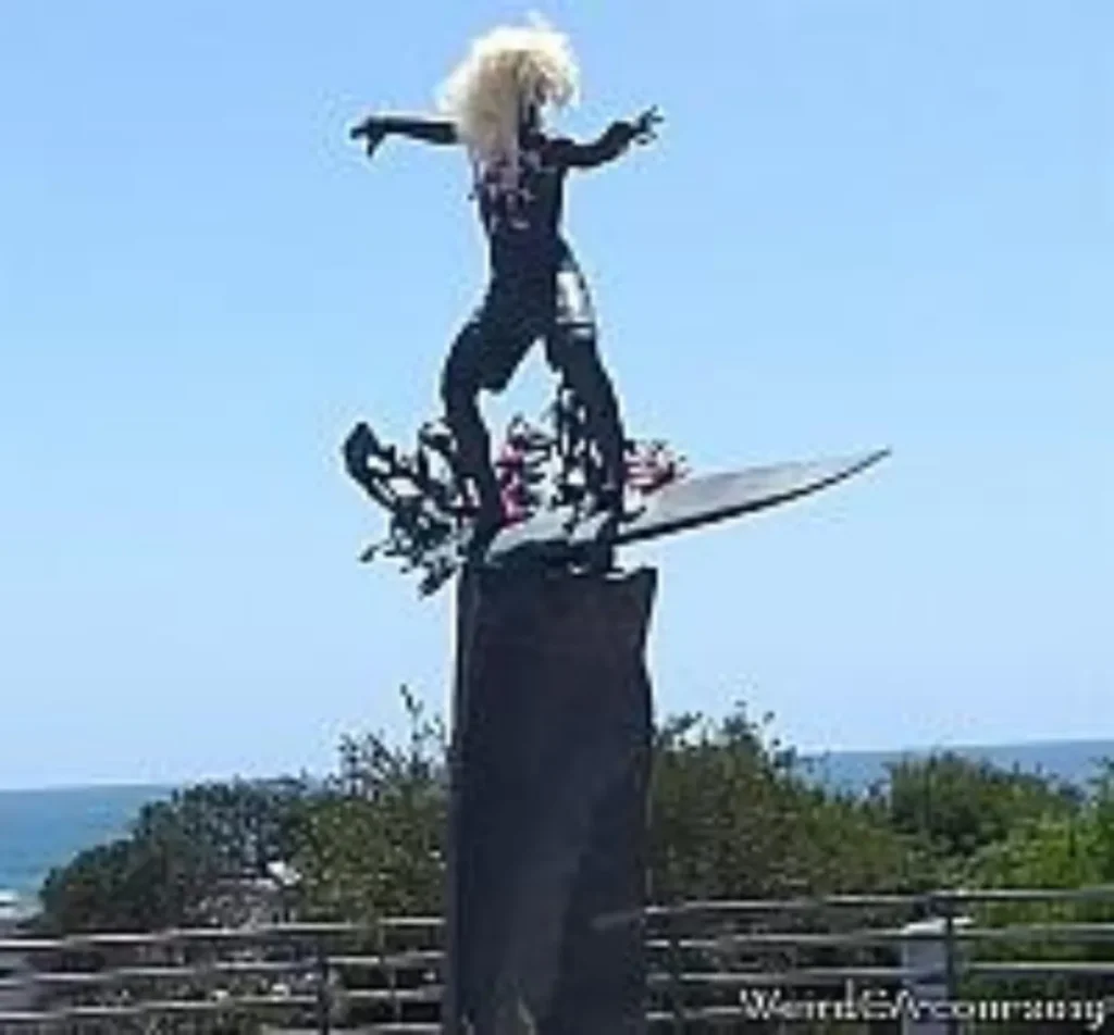 Whimsical sculpture of a surfer figure with wild hair balancing on a surfboard atop a tall pedestal outdoors.