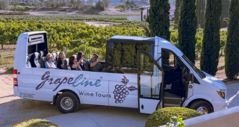 Passengers enjoying a vineyard tour aboard a Grapeline Wine Tours shuttle bus. The open-air tour vehicle is parked beside rows of grapevines and tall cypress trees in a scenic wine country setting.